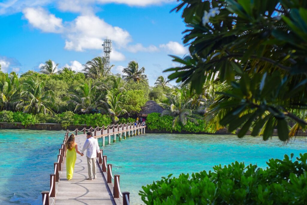 Couple holding hands on a boardwalk in paradise with turquoise water and lush greenery.